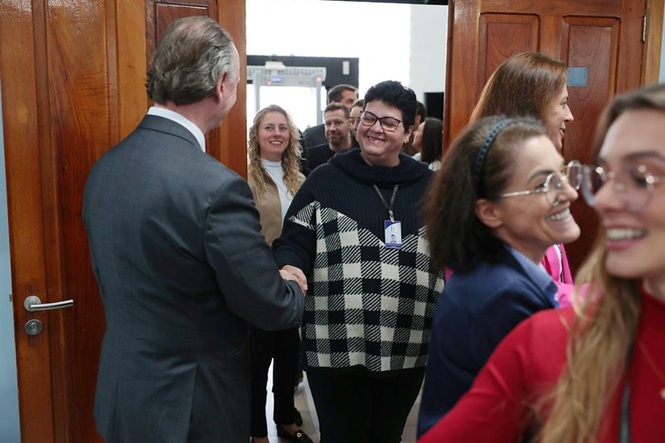 Foto horizontal: o desembargador Rubens Schulz, de costas, cumprimenta visitante em ambiente institucional, gesto de acolhimento e proximidade.
