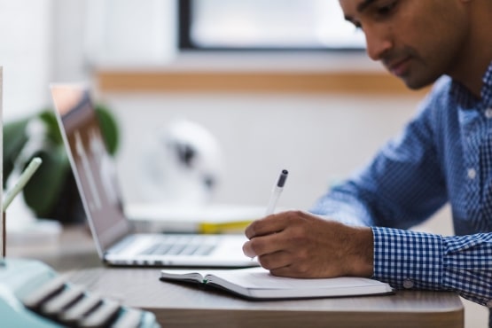 Foto horizontal: homem escreve em caderno ao lado de notebook, ambiente de estudo, clima de concentração e aprendizado.
