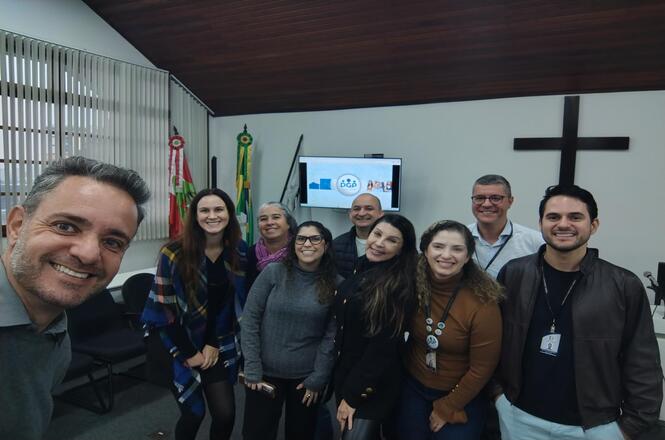 Foto horizontal: grupo sorri em registro coletivo em ambiente institucional, clima de integração e fortalecimento de vínculos.