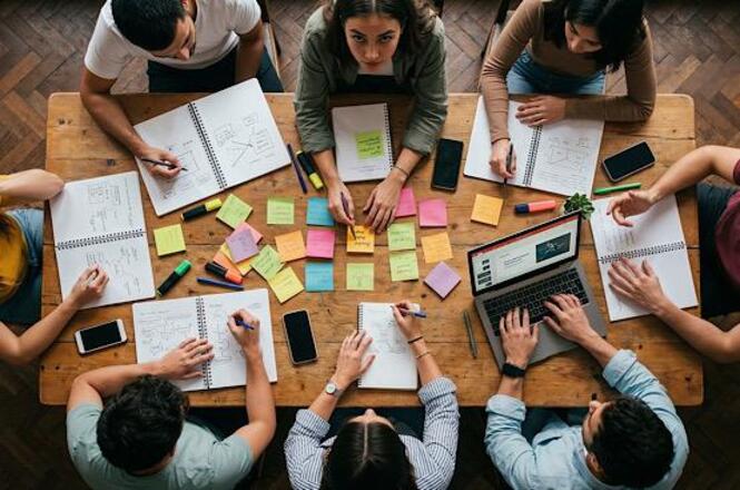 Foto horizontal: equipe colabora em mesa com anotações e post-its, planejamento coletivo em andamento, clima de organização e trabalho conjunto.