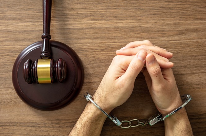 Foto horizontal: mãos algemadas sobre mesa ao lado de martelo da justiça, simbolizando responsabilização e ação judicial.