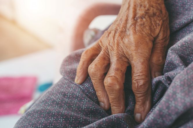 Foto horizontal: mão de pessoa idosa em destaque, textura da pele evidenciada, clima de cuidado, tempo e vulnerabilidade.
