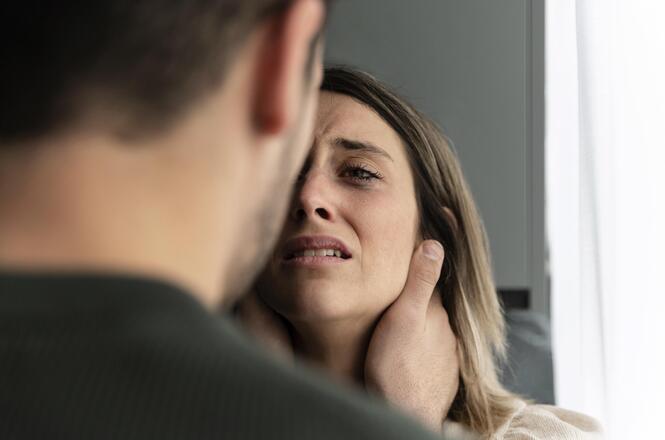 Foto horizontal: mulher com expressão de sofrimento é segurada pelo rosto, cena sugere situação de violência, clima de tensão e vulnerabilidade.