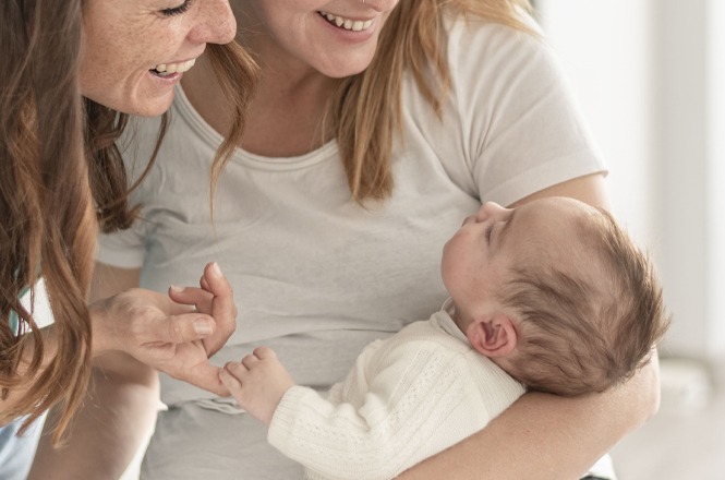 Foto horizontal: duas mulheres interagem com bebê no colo, expressão de afeto e cuidado, clima de acolhimento e vínculo familiar.