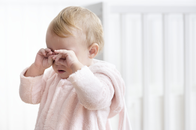 Foto horizontal: bebê com expressão de choro esfrega os olhos, ambiente interno claro, clima de fragilidade e cuidado infantil.