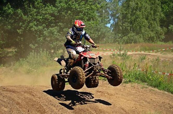 Foto horizontal: piloto salta com quadriciclo em pista de terra, poeira ao redor, clima de adrenalina e esporte off-road.