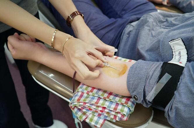 Foto horizontal: profissional realiza coleta de sangue em paciente, procedimento clínico em andamento, clima de cuidado e atendimento em saúde.