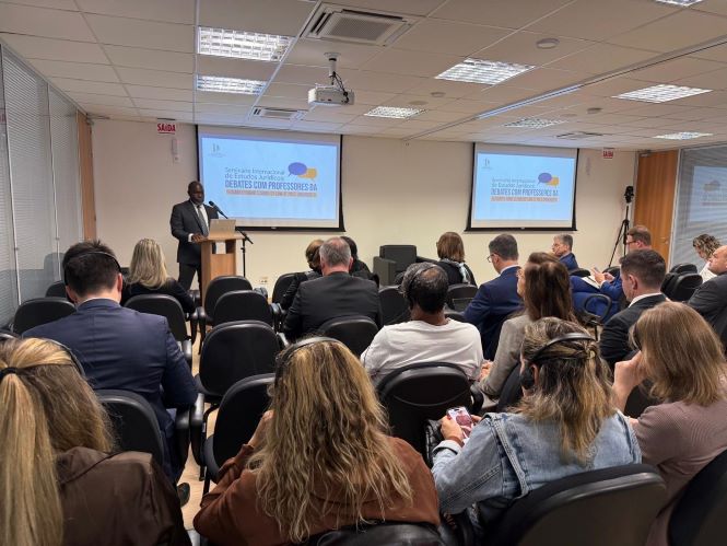 Fotografia colorida registrada em uma sala de eventos da Academia Judicial. Em primeiro plano, participantes sentados em cadeiras pretas acompanham uma palestra. Ao fundo, o reitor da Pace University, Horace Anderson Jr., um homem negro, veste terno escuro e gravata e fala ao púlpito, posicionado à frente da sala. Atrás dele, dois telões exibem a identidade visual do “Seminário Internacional de Estudos Jurídicos: Debates com Professores da Pace University”. O ambiente é iluminado, com paredes claras, teto rebaixado e equipamentos de projeção. O público, formado por homens e mulheres, observa atentamente a apresentação.