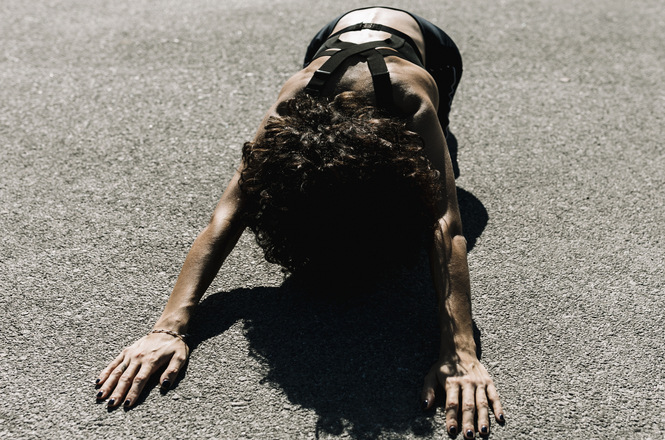 Foto horizontal: mulher ajoelhada no chão com o corpo inclinado para frente, mãos apoiadas no asfalto, clima de dor, exaustão e vulnerabilidade.