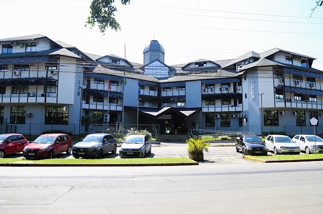 Foto horizontal: fachada do Fórum de Joinville, edifício institucional com veículos estacionados à frente, clima de acesso à Justiça e funcionamento administrativo.