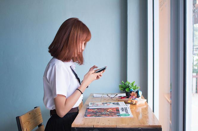 Foto horizontal: jovem utiliza celular em ambiente interno, apoiada em mesa próxima à janela, clima de concentração, cotidiano e conexão digital.