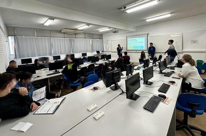 Foto horizontal: alunos acompanham aula em laboratório de informática no programa Novos Caminhos, apresentação ao fundo, clima de aprendizado, inclusão e capacitação profissional.