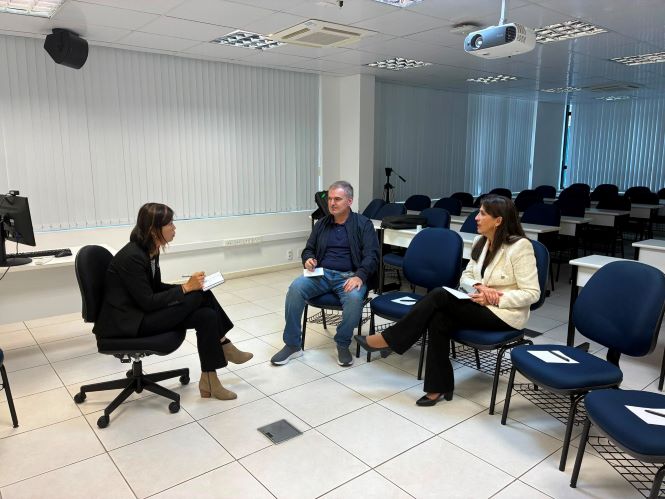 Fotografia realizada em uma sala de aula da Academia Judicial, com cadeiras azuis organizadas em fileiras ao fundo. Em primeiro plano, a professora Siyu Liu está sentada em uma cadeira, de perfil, conduzindo uma entrevista e segurando anotações. À sua frente, estão sentados o juiz Fernando Vieira Luiz, ao centro, vestindo jaqueta escura e calça jeans, e a juíza Andreia Vaz, à direita, usando blazer branco e calça preta. Os três participam de uma conversa em ambiente acadêmico, com projetor instalado no teto e iluminação clara.