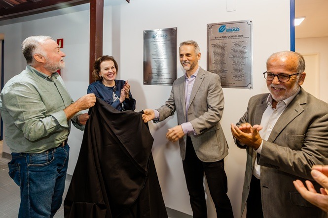 Foto horizontal: autoridades participam de descerramento de placa em ambiente institucional, gesto simbólico em destaque, clima de celebração e reconhecimento.
