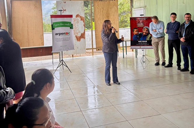 Foto horizontal: mulher fala ao público em atividade na Terra Indígena Laklãnõ, autoridades acompanham ao fundo, cartazes institucionais em destaque, clima de diálogo, inclusão e ação social.