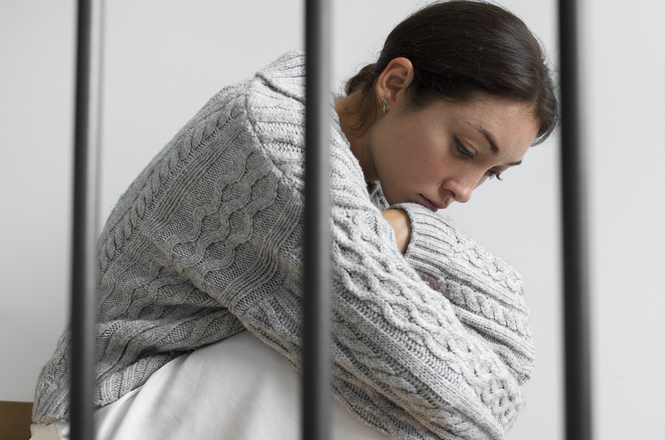 Foto horizontal: mulher sentada com postura retraída e olhar baixo, braços envoltos ao corpo, grades em primeiro plano, clima de vulnerabilidade, sofrimento e privação.