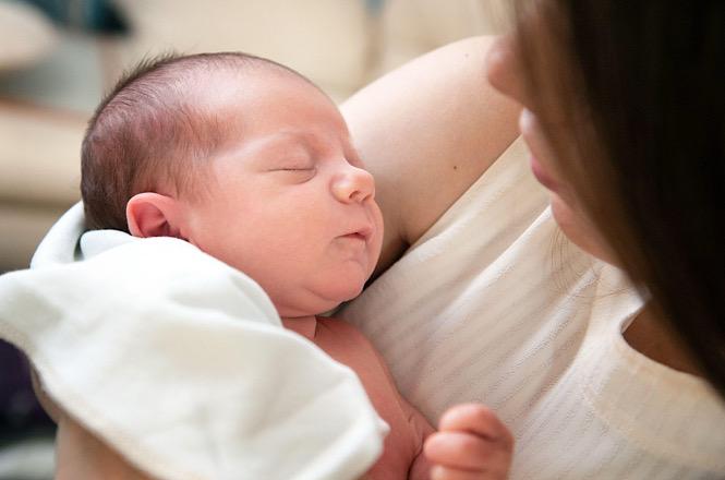 Foto horizontal: bebê dorme no colo de mulher, rosto sereno em destaque, clima de cuidado, afeto e proteção materna.