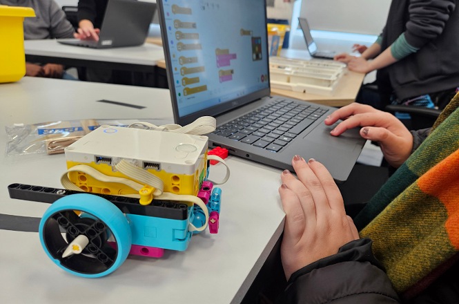 Foto horizontal: estudante utiliza notebook ao lado de robô educacional em montagem, atividade de programação em destaque, clima de inovação, aprendizado e tecnologia.