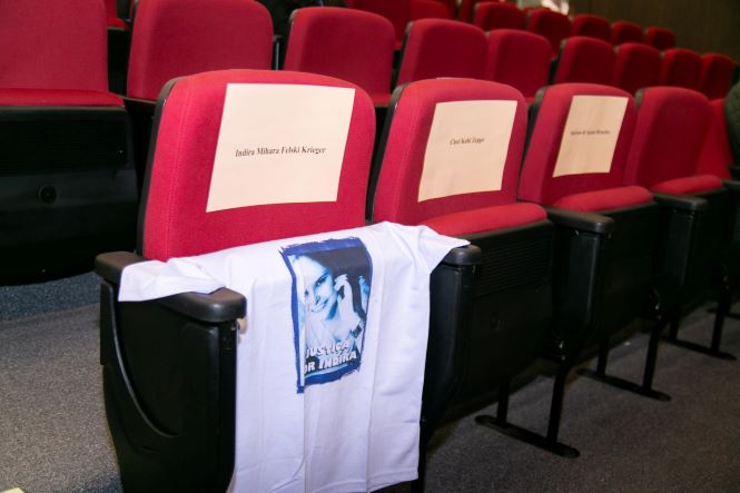 Fotografia de três poltronas vermelhas de auditório reservadas e vazias. Sobre os encostos há folhas brancas com nomes impressos. Na primeira poltrona, uma camiseta branca está estendida, trazendo a imagem de uma mulher e a inscrição “Justiça por Indira”. A cena simboliza a homenagem e a lembrança de vítimas de feminicídio, em contraste com as cadeiras vazias ao fundo do auditório.