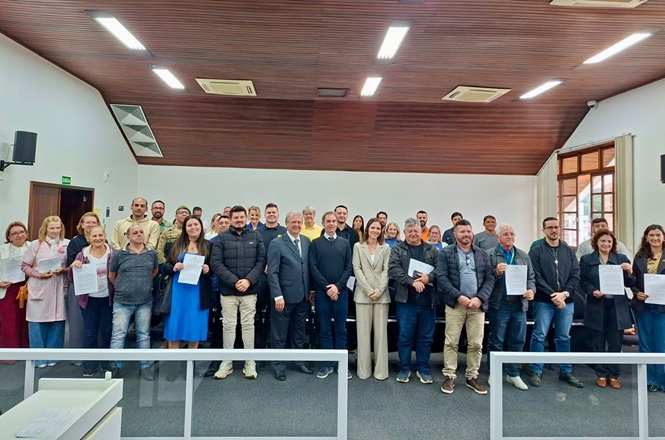 Foto horizontal: grupo de participantes posa com certificados em ambiente institucional, documentos em destaque, clima de conquista, reconhecimento e fortalecimento comunitário.