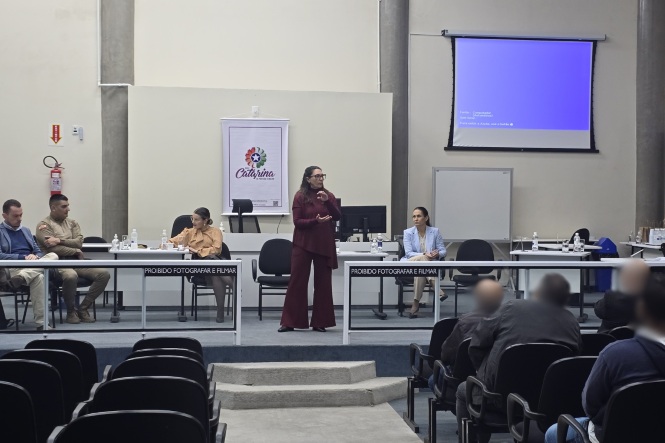 Foto horizontal: palestrante conduz apresentação em auditório com mesa de autoridades ao fundo, público presente em destaque, clima de debate, capacitação e diálogo institucional.
