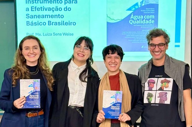 Foto horizontal: grupo de participantes posa com publicações durante atividade acadêmica sobre saneamento básico, apresentação ao fundo em destaque, clima de aprendizado, pesquisa e divulgação científica.