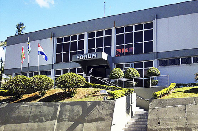 Foto horizontal: fachada de fórum com arquitetura moderna e paisagismo frontal, acesso por escadaria e rampa em destaque, clima institucional e presença do Poder Judiciário.