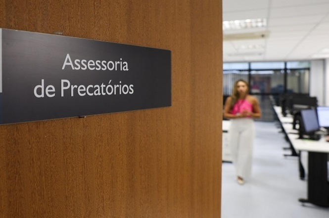 Foto horizontal: placa indica “Assessoria de Precatórios” em ambiente interno do Judiciário, setor administrativo ao fundo, clima institucional, organização e atendimento público.