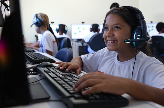 Criança usa computador com fone em sala de informática, sorrindo durante atividade digital e aprendizado tecnológico