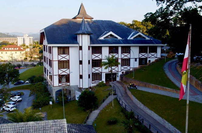Foto horizontal: edifício institucional com arquitetura em estilo enxaimel, área verde ao redor, clima de organização e presença pública