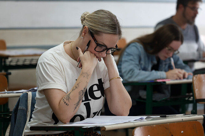 Foto horizontal: candidata concentrada realiza prova em sala de aula, com outros participantes desfocados ao fundo.