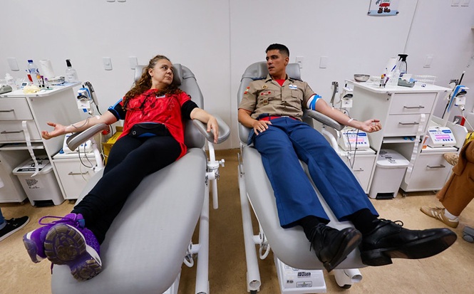 Foto horizontal: mulher e policial militar doam sangue em cadeiras reclináveis de hemocentro.