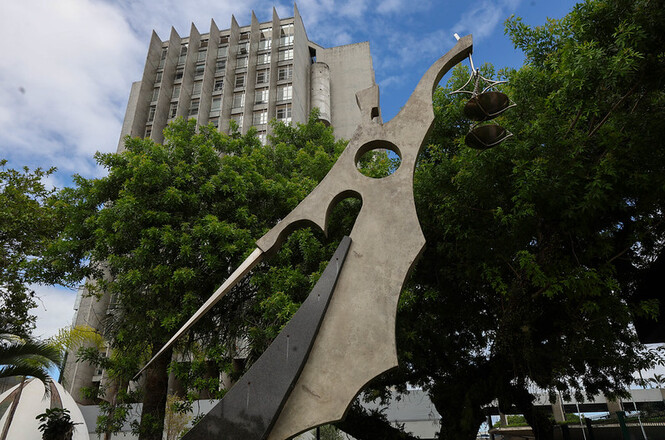 Foto horizontal: escultura da Justiça em primeiro plano diante da sede do TJSC cercada por árvores sob céu parcialmente nublado.
