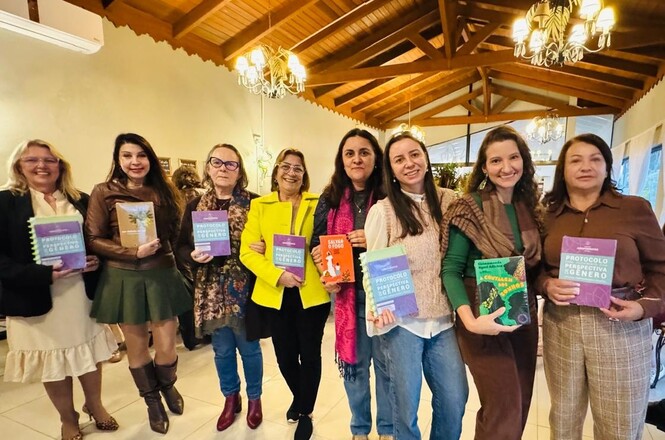 Foto horizontal: grupo de mulheres exibe publicações sobre gênero, direitos e proteção em ambiente de encontro institucional.