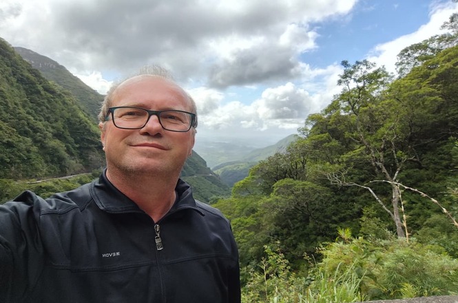 Foto horizontal: homem de óculos posa para selfie diante de vale cercado por montanhas e vegetação exuberante sob céu nublado.