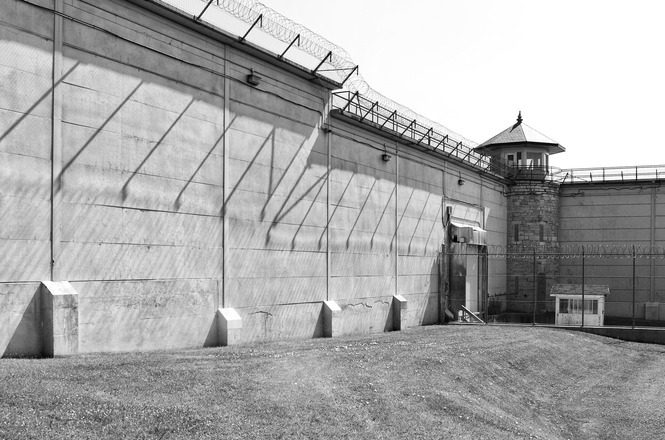 Foto horizontal em preto e branco: muralha de unidade prisional com cercas de segurança, torre de vigilância e área externa cercada por arame farpado.