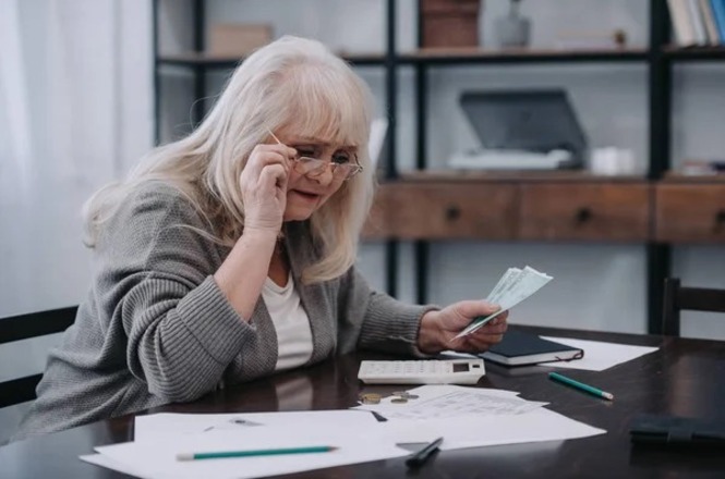 Foto horizontal: mulher idosa confere dinheiro e documentos sobre a mesa, ao lado de calculadora e anotações financeiras.