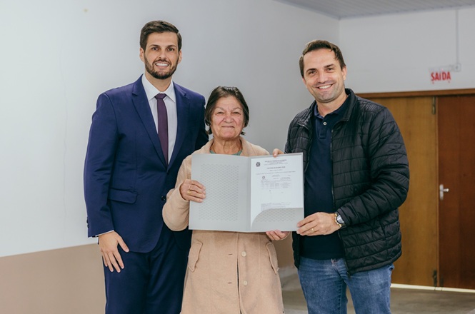 Foto horizontal: mulher idosa exibe documento ao lado de dois homens durante cerimônia de entrega em ambiente institucional.