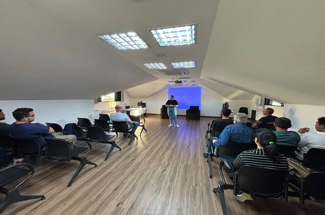 Foto horizontal: participantes acompanham atividade educativa em sala ampla, com cadeiras espaçadas e apresentação projetada ao fundo.