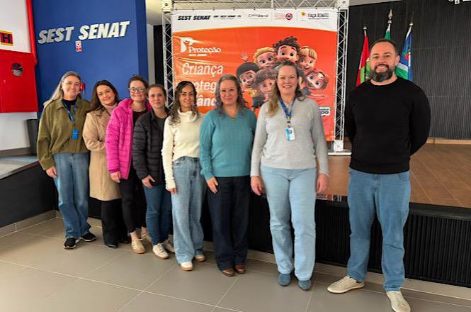 Foto horizontal: grupo de profissionais posa diante de painel sobre proteção de crianças e adolescentes em evento no Sest Senat.