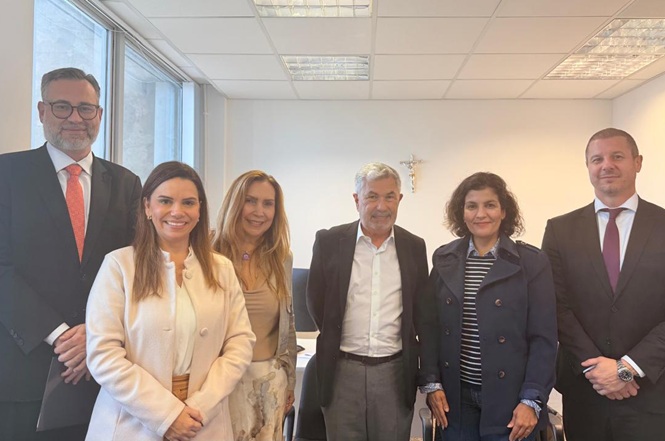 Foto horizontal: grupo de cinco pessoas posa em ambiente institucional, diante de parede clara com crucifixo ao fundo.