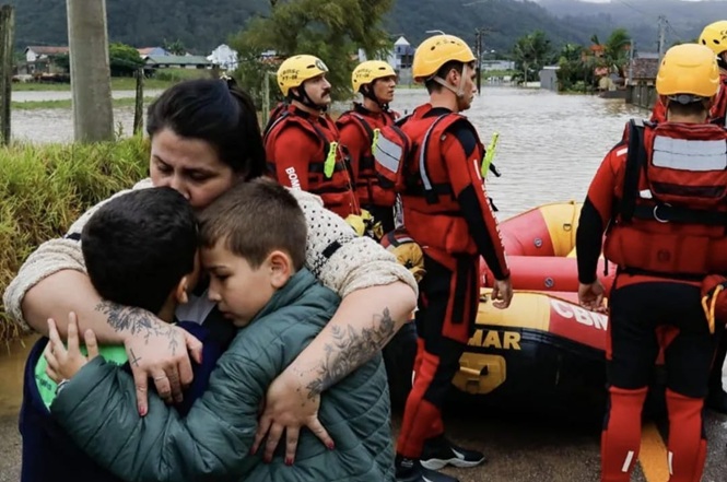 Foto horizontal: mulher abraça duas crianças em área alagada enquanto equipes de resgate atuam ao fundo com bote inflável.