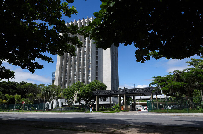 Foto horizontal: fachada da sede do Tribunal de Justiça de Santa Catarina cercada por árvores sob céu azul com nuvens.