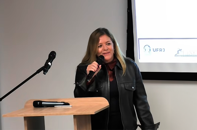 Foto horizontal: mulher fala ao microfone em evento acadêmico, ao lado de púlpito e tela de apresentação ao fundo.