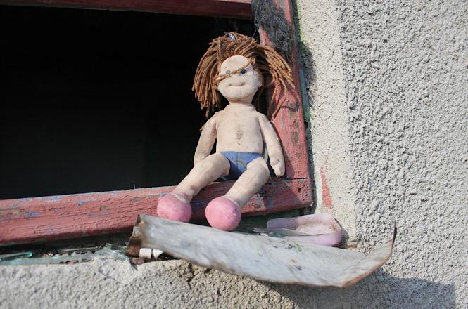 Foto horizontal: boneca desgastada repousa na janela de uma construção simples, simbolizando vulnerabilidade e infância em contexto de risco social.