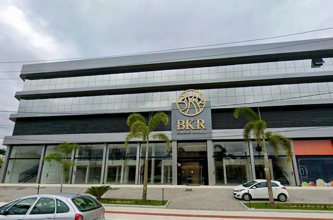 Foto horizontal: fachada do BKR Park Mall, centro comercial de arquitetura moderna com palmeiras e veículos estacionados em frente.