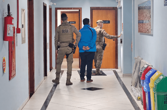 Foto horizontal: policiais militares conduzem pessoa algemada por corredor de unidade pública, em cumprimento de procedimento policial.
