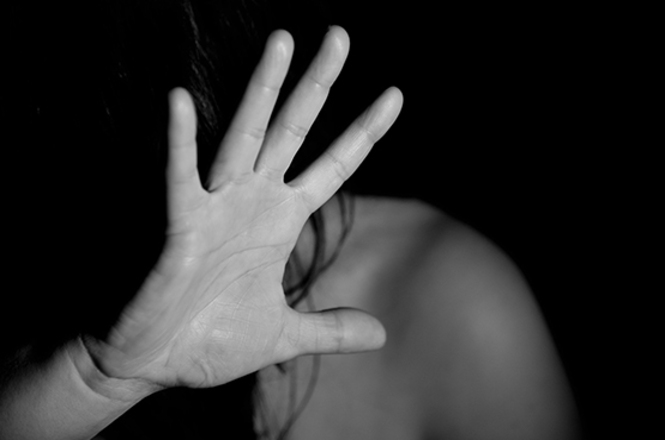 Foto ilustrativa em preto e branco: mulher ergue a mão em sinal de proteção e recusa, simbolizando violência, medo e pedido de ajuda.
