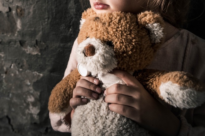 Foto ilustrativa: criança abraça um urso de pelúcia em ambiente escuro, simbolizando vulnerabilidade, proteção e os direitos da infância.