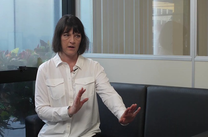Foto horizontal: mulher concede entrevista em ambiente institucional, gesticulando durante conversa registrada em vídeo.